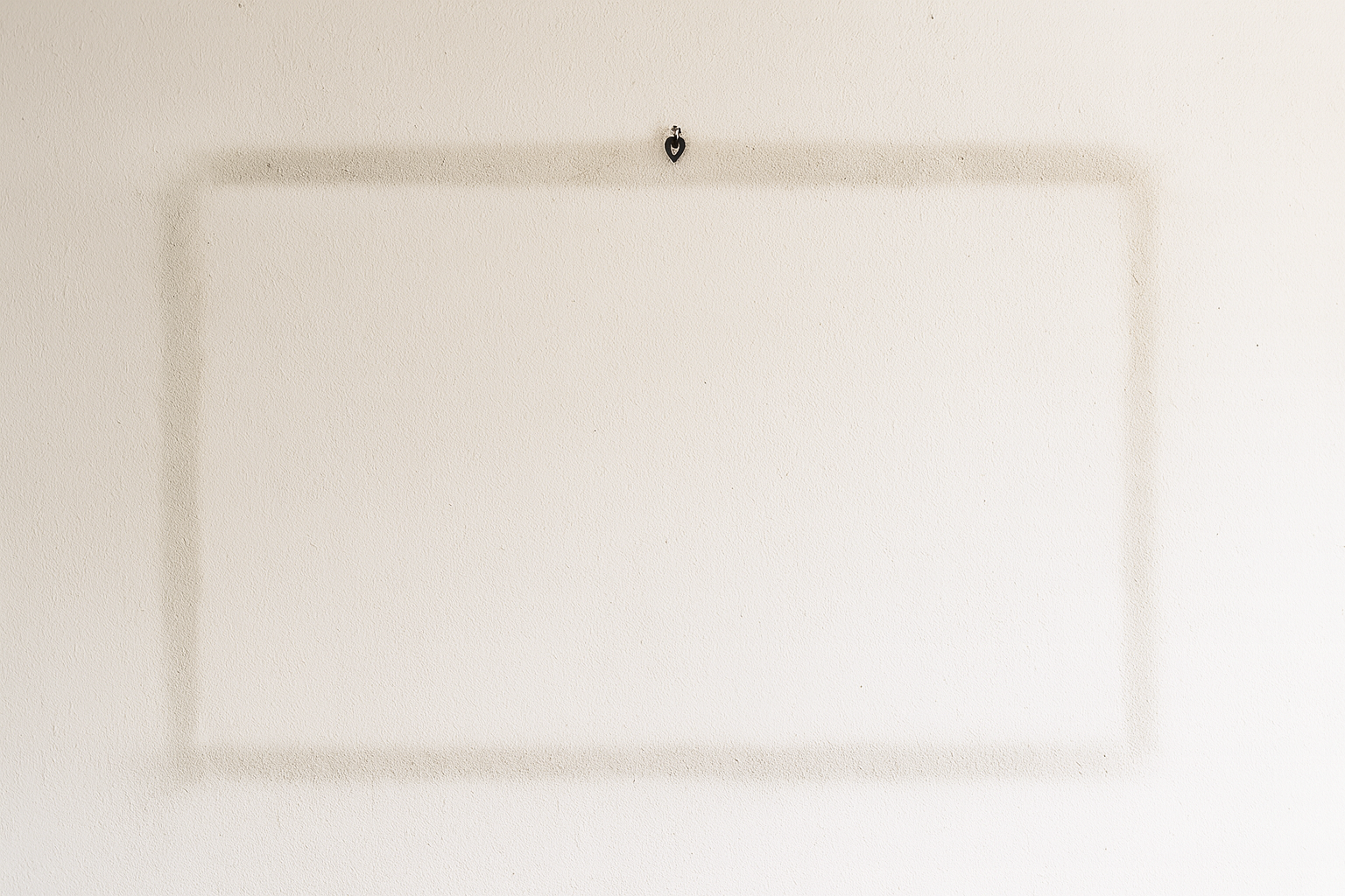 A plain off-white wall showing a faint rectangular outline where a picture frame once hung, with a single metal hook still in place.
