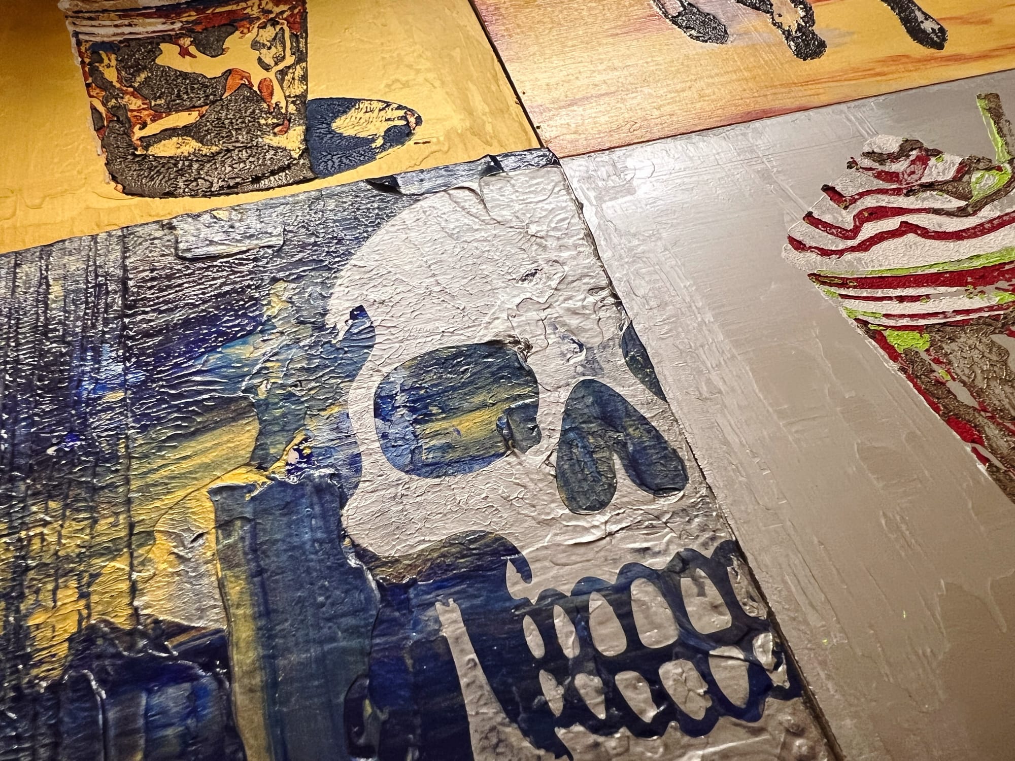 Close-up of textured acrylic paintings showing a skull, a coffee cup, and a whipped-cream drink, with shimmering metallic layers catching the light. 